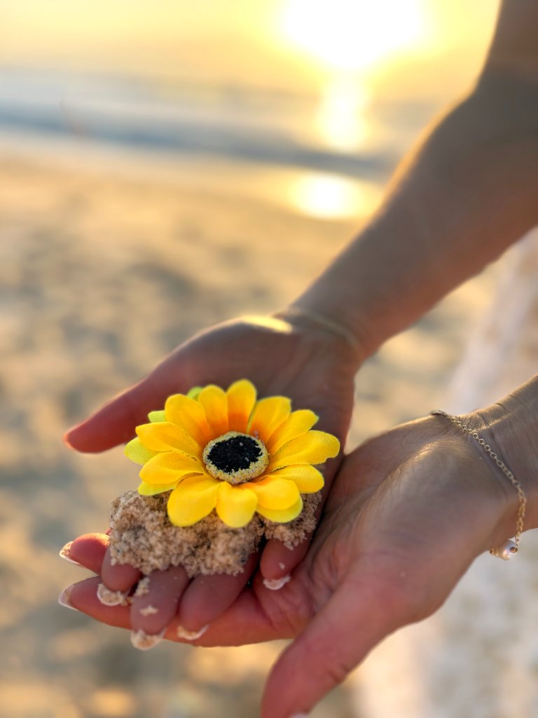 Hands holding a bright yellow blossom in sand with sun flare